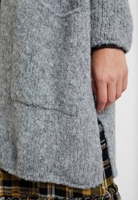 Gray, textured knit coat with a relaxed fit, featuring side slits and a visible pocket, worn over a black and yellow checkered skirt.