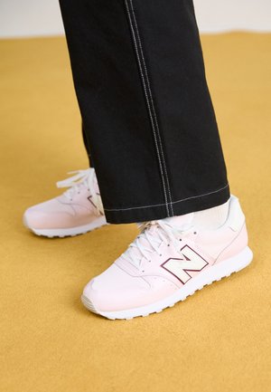 Osoba ubrana w czarne spodnie i jasnoróżowe buty New Balance stoi na podłodze w kolorze musztardowej żółci.