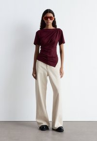 Bordeaux-farbenes, kurzärmliges Oberteil mit drapierten Details, kombiniert mit cremefarbenen, weit geschnittenen Hosen und schwarzen Plateauschuhen. Das Model trägt orangefarbene Sonnenbrillen.