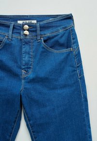 Blue denim jeans with double metal buttons, belt loops, front and side pockets, and visible stitching details.