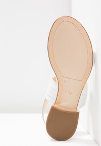 Sandale à talon blanche appuyée contre un mur, montrant la semelle beige avec une semelle antidérapante en caoutchouc et la marque "hög | taille 5".