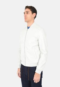 Giacca bomber bianca in pelle con chiusura a zip, polsini e vita a coste. Presenta tasche laterali e una texture liscia. Indossata con jeans scuri.