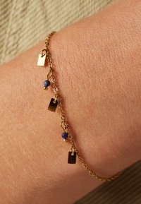 Gouden kettingarmband met kleine rechthoekige bedels en blauwe kralen, voorzien van een gladde textuur en fijne schakels, gedragen om een pols.