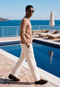 Homme en lunettes de soleil marchant à côté d'une piscine avec des chaises longues blanches et un parasol, surplombant une mer bleue avec des voiliers sous un ciel dégagé.