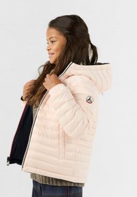 Giacca puffer rosa chiaro con cappuccio, chiusura a zip e texture a coste. Presenta un tessuto nero all'interno e una toppa con logo sulla manica.