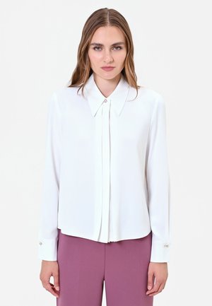 Donna con lunghi capelli mossi che indossa una camicetta bianca con bottoni e pantaloni viola, in piedi davanti a uno sfondo chiaro e uniforme.
