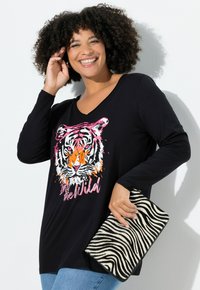 Schwarzes Langarmoberteil mit einem bunten Tiger-Graphic und dem Text "In the Wild". Das Modell hält eine Tasche mit Zebra-Muster.