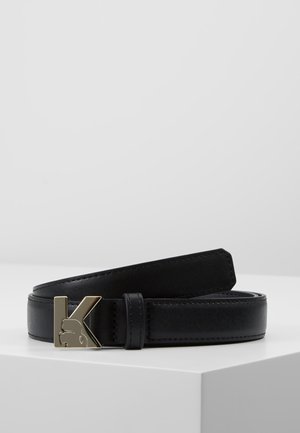 Zwarte leren riem met een gouden metalen gesp in de vorm van een "K" met een gestileerd gezichtsprofiel, opgerold weergegeven op een witte ondergrond.