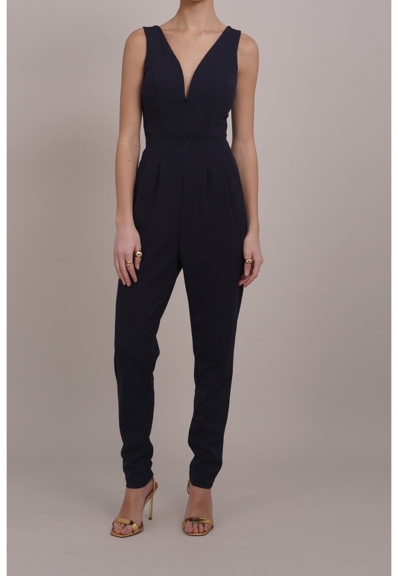 Granatowy, bezrękawnikowy kombinezon z głębokim dekoltem w kształcie V, dopasowany krój, zwężane nogawki i subtelne zaszewki. Połączony ze złotymi sandałkami na wysokim obcasie.