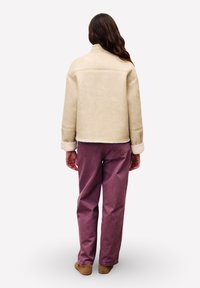 Persoon met lang zwart haar, gekleed in een beige schapenvachtjacket, paarse spijkerbroek en bruine schoenen, staand met de rug naar de camera op een witte achtergrond.