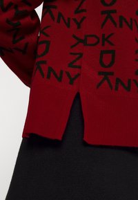 Roter gestrickter Pullover mit schwarzem Logo-Muster, gerippten Bündchen und Saum sowie seitlichen Schlitzen für mehr Bewegungsfreiheit.