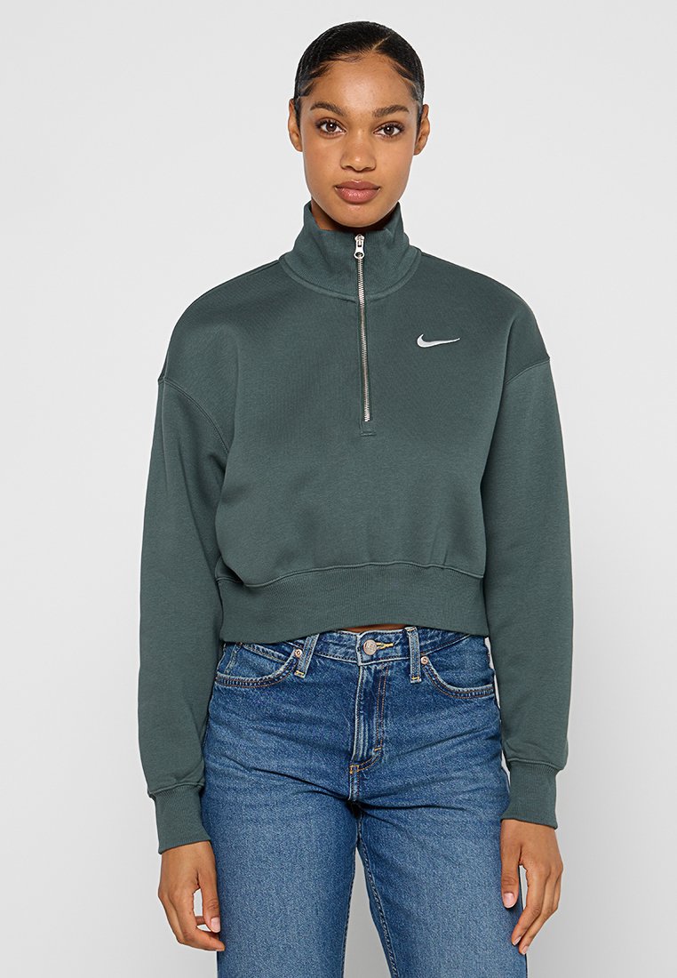 Sudadera verde corta con cuello alto y media cremallera. Presenta un logo de Nike en plata en el pecho y puños acanalados. Combinada con jeans azules.
