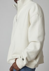 Sudadera de cremallera corta en color crema, hecha de un tejido suave. Presenta puños acanalados, un corte relajado y un pequeño logo en la manga izquierda.