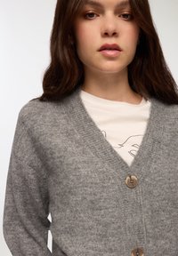 Szary sweter z dzianiny z dekoltem w serek, posiadający fakturowane wykończenie i trzy duże okrągłe guziki z przodu. Nosi się go na jasnej koszulce.