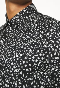 Camicetta nera con un motivo a zig zag bianco, dotata di colletto standard e bottoni. Il materiale sembra liscio con un design testurizzato.