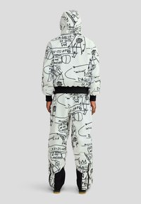 Személy fehér kapucnis pulóverben és nadrágban, melyeken fekete absztrakt vonalrajzok és szövegek láthatók, hátulról nézve, egyszínű háttér előtt.