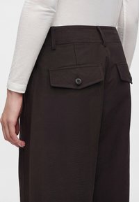 Personne portant un pantalon marron foncé avec des poches à rabat boutonnées à l’arrière et un haut blanc à manches longues, visible de la taille jusqu’en bas.