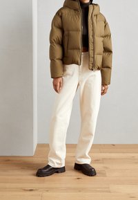 Olivgrön pufferjacka med en kortad design, som har dragkedja fram, stora fickor och huva. Matchad med off-white vida jeans och bruna skor.