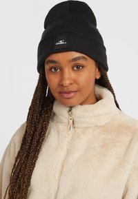 Zwarte geribbelde gebreide beanie met een vierkante logopatch, gedragen met een beige fleece jack met rits en hoge kraag.