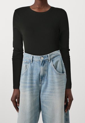 Persoon draagt een zwarte, nauwsluitende top met lange mouwen, ingestopt in een lichtblauwe, wijd uitlopende jeans met hoge taille en voorzakken.