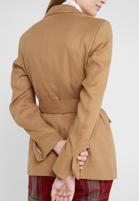 Karmelowy żakiet o dopasowanym kroju, z wysmuklającą talią, długimi rękawami i delikatnym rozkloszowaniem przy mankietach. Gładka tekstura, minimalne detale.