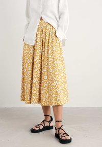 Gul kjol med vitt blommönster, plisserad design och lös passform, i kombination med svarta sandaler med remmar och en vit skjorta med knappar.