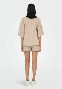 Beige Kurzarmoberteil mit entspannter Passform und gerafftem Rücken. Kombiniert mit passenden beigen Shorts und weißen Slippern.