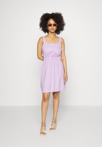 Vestido sem mangas em lavanda com cintura elástica franzida, decote quadrado e alças com volantes. Combinado com sandálias brancas de tiras.