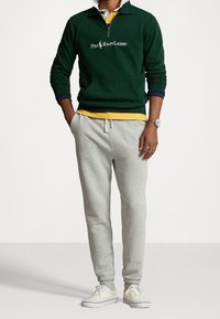 Pullover in maglia verde con colletto zip e logo, indossato sopra camicie gialle e bianche. Pantaloni della tuta grigi e scarpe beige completano l'outfit.