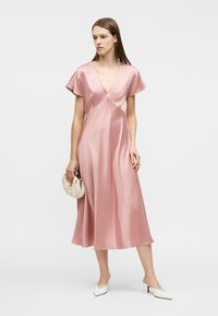 Γυναίκα που φοράει ροζ σατέν μίντι φόρεμα με κοντά μανίκια, κρατώντας μια μπεζ τσάντα φάκελο και φοράει λευκά γοβάκια με μυτερή μύτη και χαμηλό τακούνι.