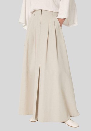 Femme portant une jupe longue beige plissée taille haute avec poches latérales et chaussures plates blanches sur un fond neutre.