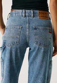 Svetlo modri jeans hlače z ohlapnim krojem, ki imajo velike zadnje žepke, usnjeno oznako in rdeče blagovne oznake. Vidne podrobnosti šivanja.