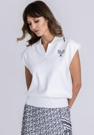 Femme portant un haut blanc sans manches avec un petit logo de raquette de tennis et une jupe plissée à motifs, regardant vers le bas avec les mains derrière le dos.