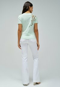 Camiseta de un suave verde menta con detalles de encaje floral en el hombro, combinada con pantalones blancos de campana, que exhiben un diseño ajustado y una textura suave.