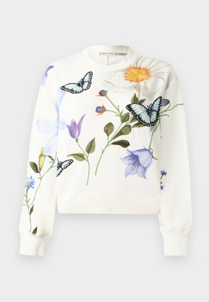 Vrouw staat met hand op heup, draagt een witte trui met bloemen- en vlinderprints, donkere wijde jeans en witte hakken.