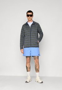 Casaco para homem com fecho éclair em azul escuro com padrão geométrico; combinado com calções azul claro e ténis brancos. Óculos de sol completam o visual.