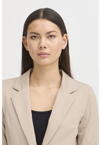 Junge Frau mit glattem braunem Haar, die einen beigen Blazer über einem schwarzen Oberteil trägt und mit neutralem Gesichtsausdruck geradeaus blickt.