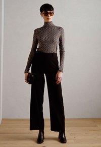 Pullover mit hohem Kragen in einem welligen Schwarz-Weiß-Muster, kombiniert mit hochtaillierten schwarzen Weitbein-Hosen und schwarzen Ankle Boots. Schwarze Sonnenbrille und Clutch.
