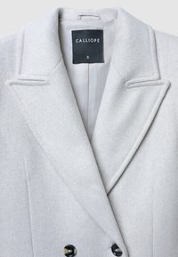 Cappotto in lana grigio chiaro a doppiopetto con ampi risvolti e bottoni neri, che mostra l'etichetta interna con il nome del marchio e la taglia piccola.