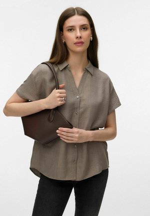 Kobieta ubrana w beżową, krótkim rękawem koszulę na guziki i czarne spodnie, trzymająca ciemnobrązową skórzaną torebkę przewieszoną przez ramię.