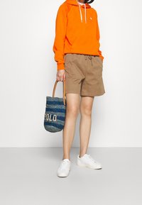 Světlě oranžová mikina s kapucí, béžové šortky, bílé tenisky a modrá tkaná taška s nápisem "POLD". Jednoduchý, ležérní outfit.