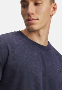 Junger Mann in einem verblassten dunkelblauen Rundhalsausschnitt-T-Shirt mit einem kleinen schwarz-weißen Logo, der nach unten und zur Seite schaut.