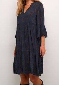 Vestido azul con escote en V y mangas cortas, con un diseño en capas y sutiles patrones marrones. Hecho de una tela ligera.