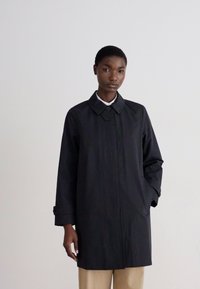 Trench de lluvia negro, hasta las rodillas, hecho de una tela suave y resistente al agua. Presenta un collar puntiagudo, cierre de botones y dos bolsillos laterales.