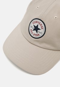 Cappellino da baseball beige in cotone con visiera arrotondata, caratterizzato da una patch circolare in bianco e nero con una stella e testo rosso.