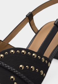 Μαύρο δερμάτινο σανδάλι με χρυσά καρφιά, πλεκτή διακόσμηση, ανοιχτή φτέρνα και ταμπά εσωτερική σόλα, εμφανιζόμενο από κοντινή γωνία.