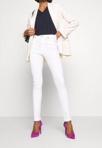 Bílé úzké džíny, černé triko s výstřihem do V, krémový blejzr a fuchsiové lodičky na vysokém podpatku. Blejzr má formální střih a knoflíky.