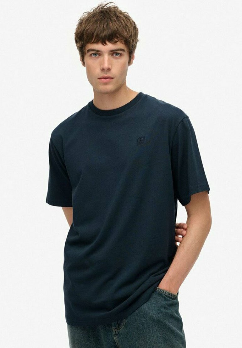 Jongeman met kort bruin haar, gekleed in een eenvoudige donkerblauwe T-shirt en blauwe jeans, staand met één hand in de zak.