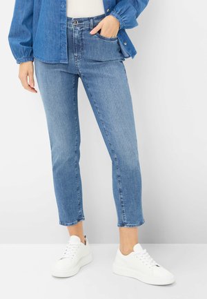 Kvinde iført blå jeans med nålestriber, en blå denimskjorte og hvide sneakers, stående med den ene hånd i lommen på en hvid baggrund.