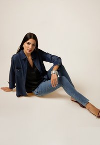 Camisa de denim en azul oscuro con botones en la parte delantera, combinada con una blusa negra y jeans skinny azul claro. Los accesorios incluyen pulseras de plata y tacones.